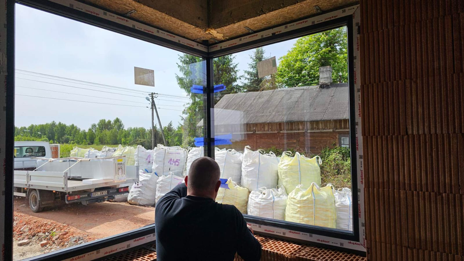Montaż narożnego okna z czarną ramą w surowym murze. Widok na podwórze z workami i samochodem dostawczym. Taśmy zabezpieczające na szybach.
