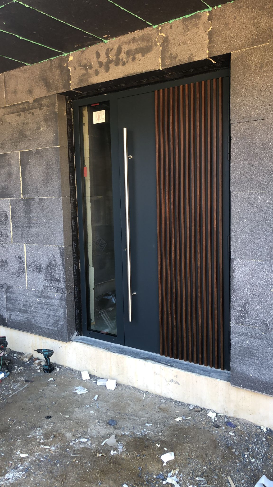 Nowoczesne drzwi zewnętrzne w kolorze grafitowym z drewnianymi lamelami i długim, srebrnym uchwytem, zamontowane w surowym murze. Widoczny wiertarko-wkrętak.