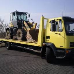 Żółta laweta DAF przewozi na platformie czarno-żółtą ładowarkę kołową Komatsu na tle brukowanej nawierzchni i drzew bez liści.