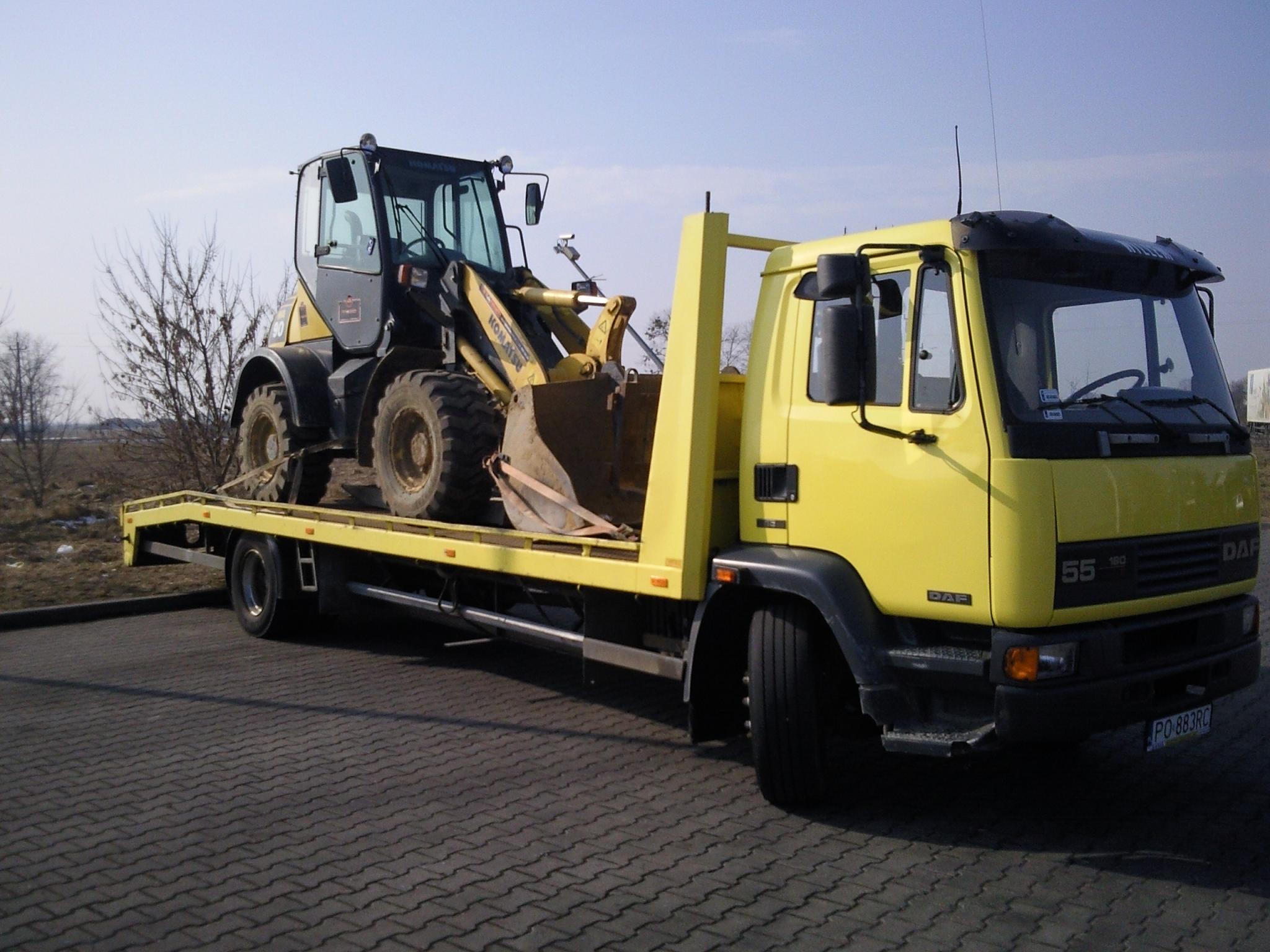 Żółta laweta DAF przewozi na platformie czarno-żółtą ładowarkę kołową Komatsu na tle brukowanej nawierzchni i drzew bez liści.