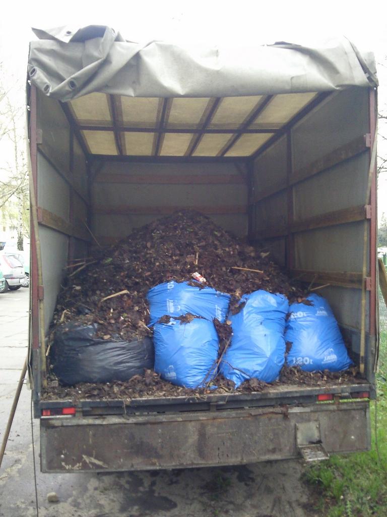 Otwarta paka ciężarówki wypełniona stertą liści i gałęzi, częściowo zapakowanych w niebieskie i czarne worki foliowe.