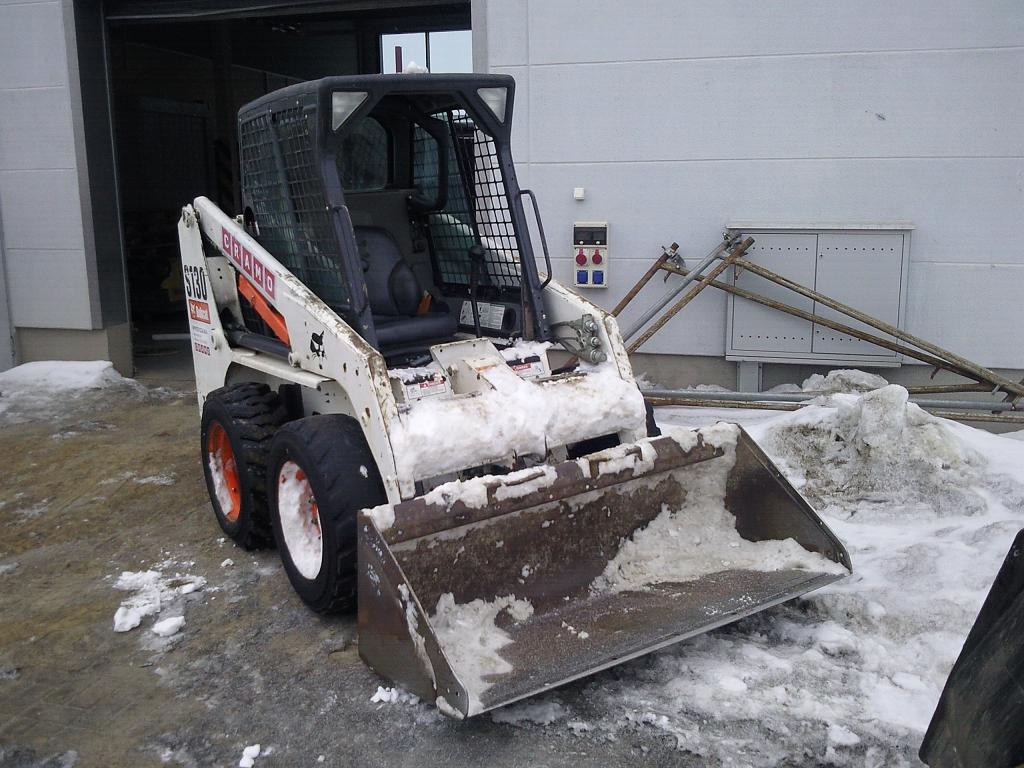 Kompaktowa ładowarka Bobcat S130 z łyżką, pokryta śniegiem, na tle szarego budynku i fragmentów rusztowania.