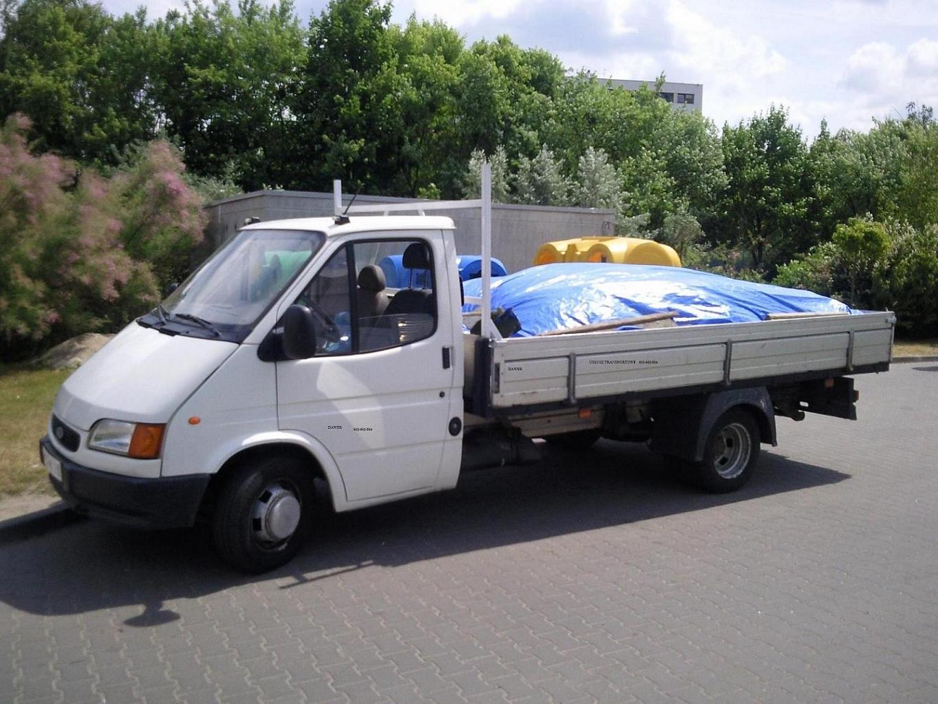 Biały samochód dostawczy z otwartą skrzynią ładunkową, zabezpieczoną niebieską plandeką, stojący na parkingu w słoneczny dzień. W tle widoczne drzewa i budynek.