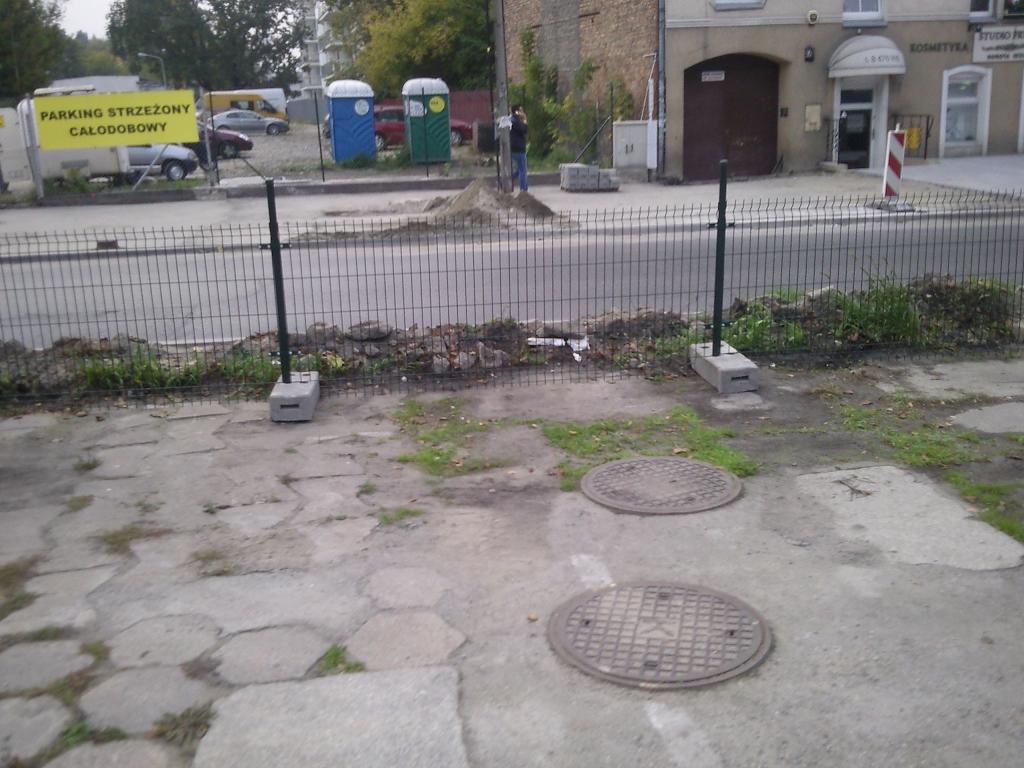 Montaż tymczasowego ogrodzenia z siatki na betonowych podstawach wzdłuż chodnika i ulicy. W tle widoczne przenośne toalety i budynek z szyldem 'Kosmetyka'.