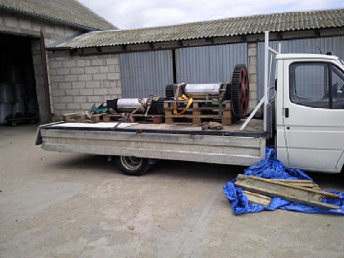 Biały samochód dostawczy z otwartą platformą przewozi na paletach elementy maszyny z kołem zamachowym na tle magazynu.