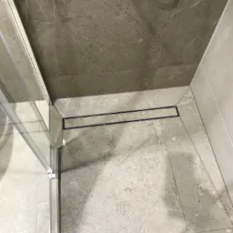 Wykończona kabina prysznicowa z odpływem liniowym i marmurowymi płytkami w odcieniach szarości. Widok z góry, fragment szklanej ściany kabiny.