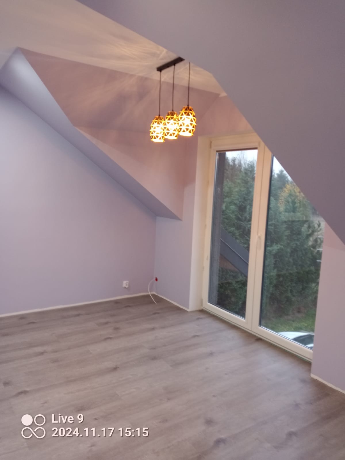 Poddasze po remoncie: fioletowe ściany, panele podłogowe, okno z widokiem na zieleń, designerskie lampy wiszące z geometrycznym wzorem.