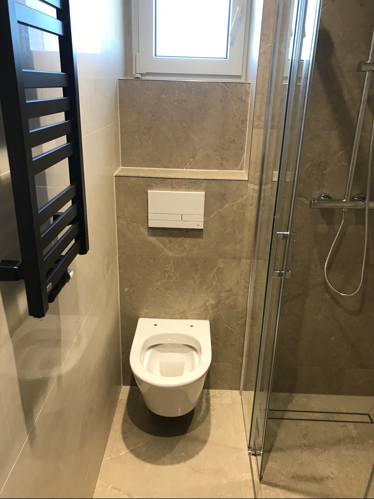 Nowoczesna łazienka po remoncie: beżowe płytki imitujące marmur, podwieszana toaleta, czarny grzejnik drabinkowy i szklana kabina prysznicowa.