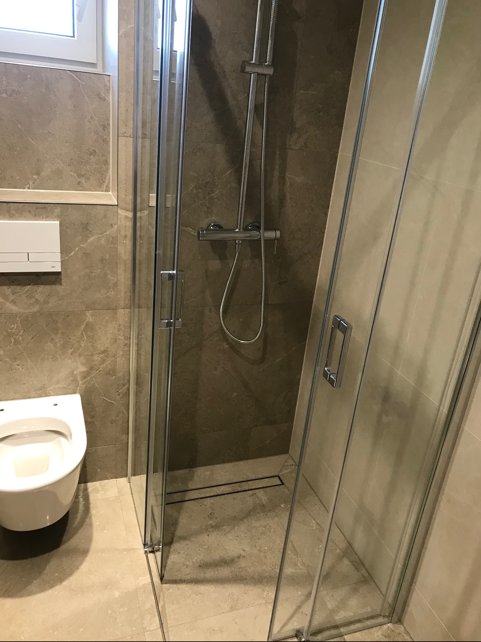 Elegancka kabina prysznicowa z deszczownicą i odpływem liniowym, widok na toaletę i beżowe płytki imitujące marmur, nowoczesna aranżacja łazienki.