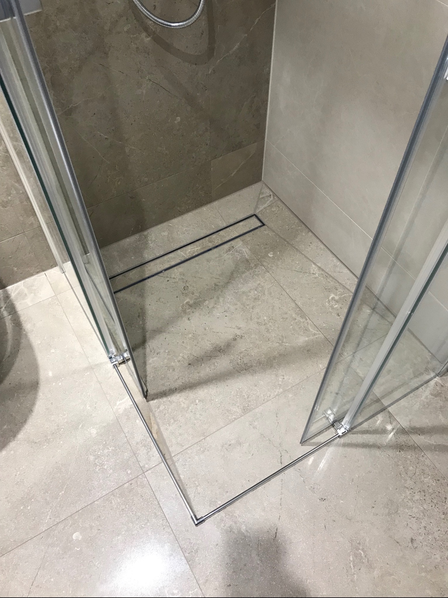 Nowoczesny prysznic walk-in z szarymi płytkami imitującymi kamień. Czarny odpływ liniowy i szklane drzwi kabiny. Minimalistyczny design.