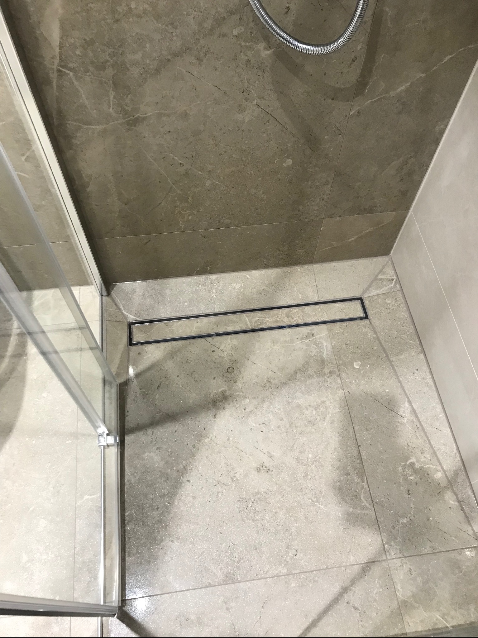 Wykończona kabina prysznicowa z odpływem liniowym i marmurowymi płytkami w odcieniach szarości. Widok z góry, fragment szklanej ściany kabiny.