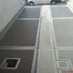 Świeżo wylana, podzielona na sekcje, betonowa posadzka z rowkowaną powierzchnią, widoczny odpływ liniowy i częściowo zaparkowany biały bus w tle.