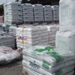 Stosy palet z workami nawozów Grupa Azoty Polifoska, widoczne etykiety i nazwy na workach, w tle budynek z napisem Węgiel Nawozy i drzewa.