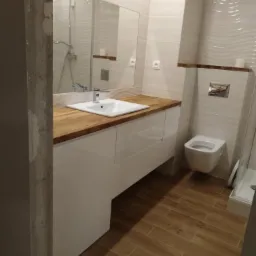 Nowoczesna łazienka z białymi szafkami pod umywalkę z drewnianym blatem, wiszącą toaletą i kabiną prysznicową w tle. Podłoga wyłożona płytkami imitującymi drewno.