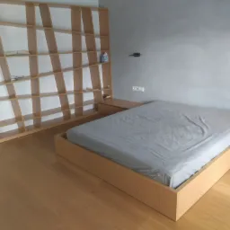 Nowoczesna sypialnia z łóżkiem na drewnianej platformie i asymetrycznym regałem z jasnego drewna.