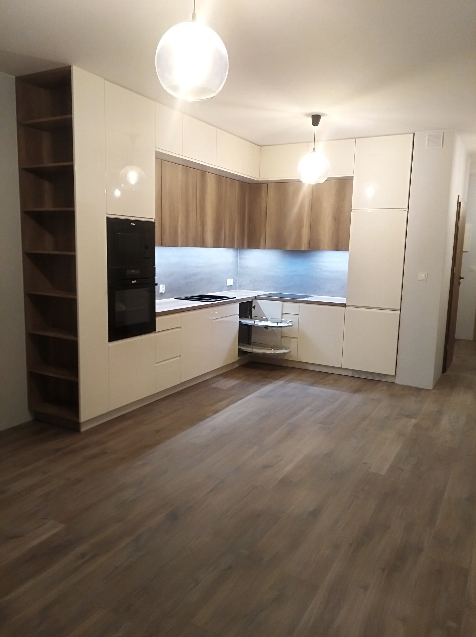 Jasna, nowoczesna kuchnia z drewnianymi akcentami, oświetlona kulistymi lampami, z widoczną szafką narożną z obrotowymi półkami i wysoką szafką z otwartymi półkami z boku.