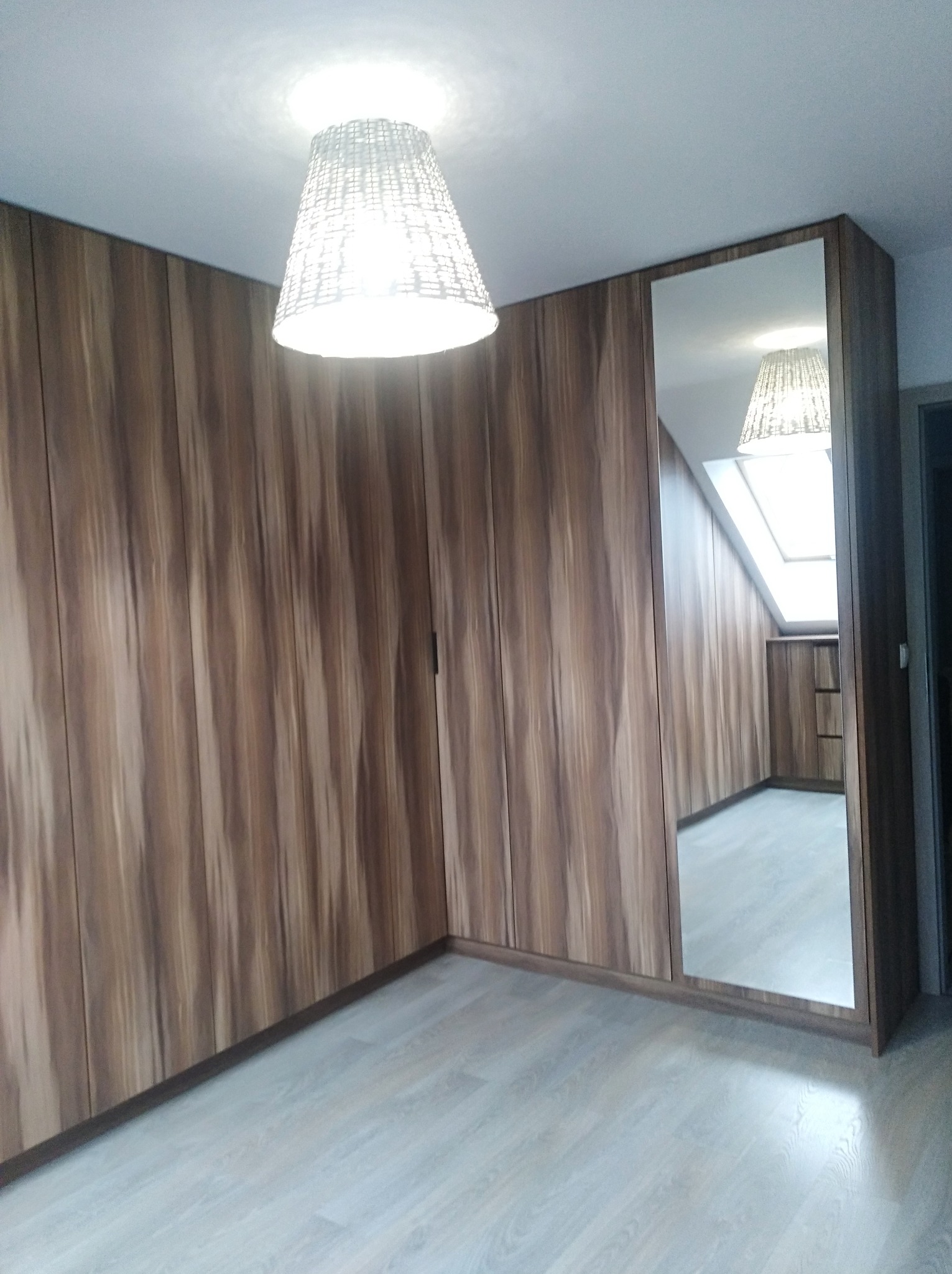 Szafa wnękowa z lustrem na całej wysokości drzwi, z drewnopodobnymi frontami w odcieniach brązu, oświetlona lampą wiszącą z abażurem z plecionki.