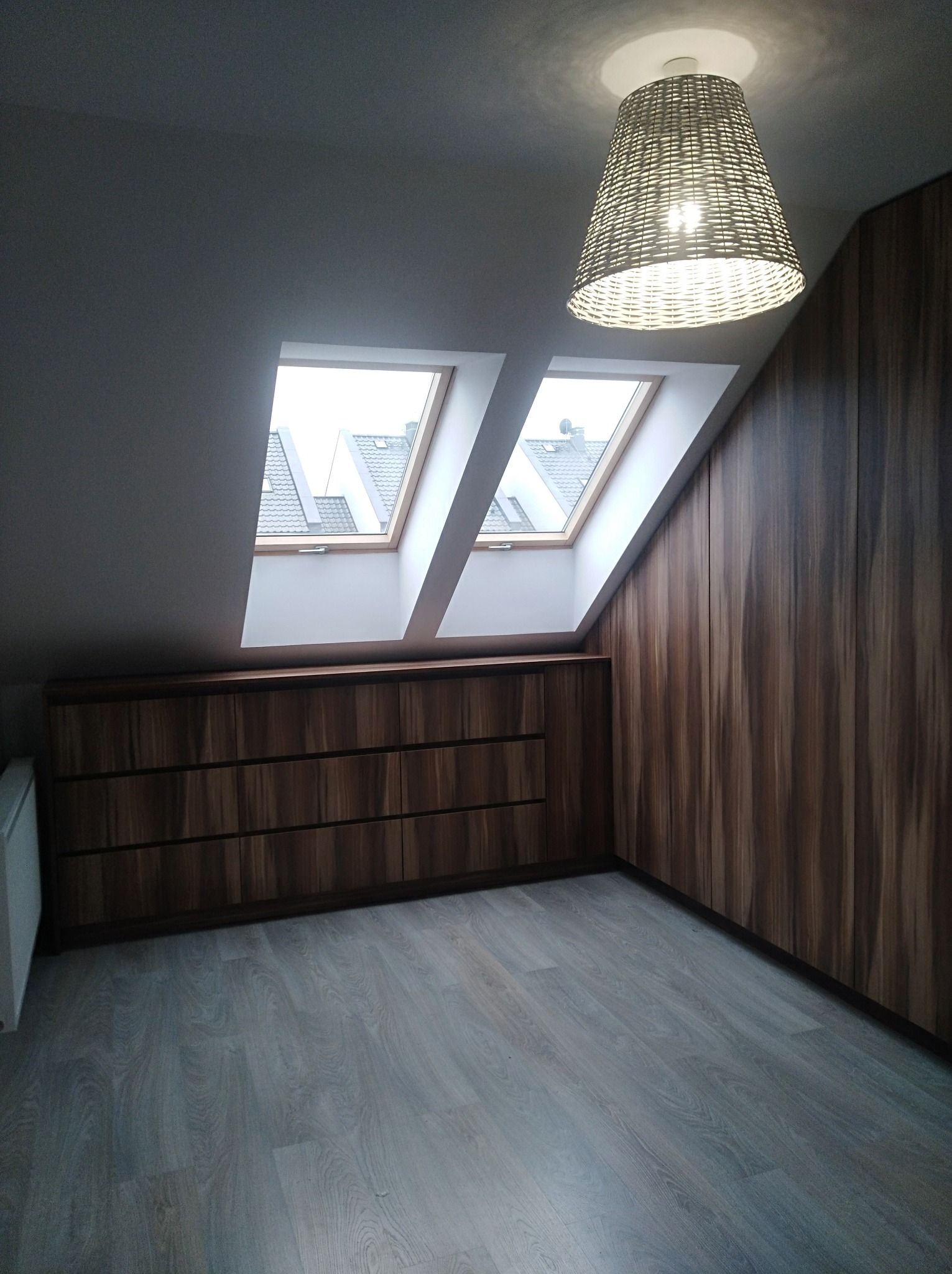 Pokój na poddaszu z zabudową meblową z forniru w odcieniach brązu, obejmującą komodę z szufladami i wysoką szafę wnękową, oświetlony lampą z wikliny i światłem z dwóch okien dachowych.