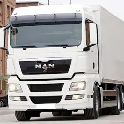 Biała ciężar&oacute;wka MAN z naczepą na tle miejskiej zabudowy z cegły. Widoczny fragment chodnika i jezdni.