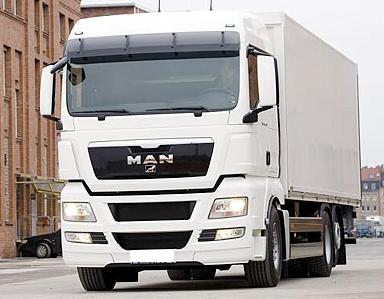 Biała ciężarówka MAN z naczepą na tle miejskiej zabudowy z cegły. Widoczny fragment chodnika i jezdni.