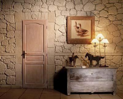 Ściana wykończona płytkami kamieniopodobnymi imitującymi piaskowiec, z drewnianą ramą drzwi po lewej i komodą z dekoracjami (lampy, obrazek, figurki koni) po prawej.