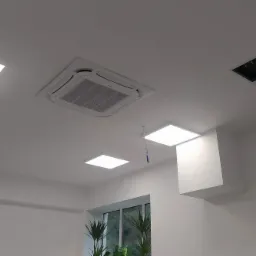 Kasetonowa jednostka klimatyzacji zamontowana w suficie podwieszanym biura, widoczne również panele oświetleniowe LED i fragment okna z roślinami.