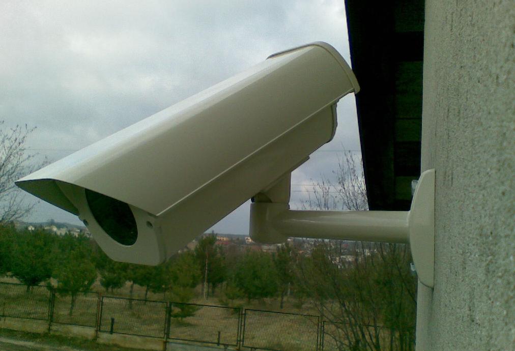 Zewnętrzna kamera monitoringu w obudowie ochronnej, zamontowana na elewacji budynku z widokiem na zieleń i zabudowania w tle, pochmurne niebo.