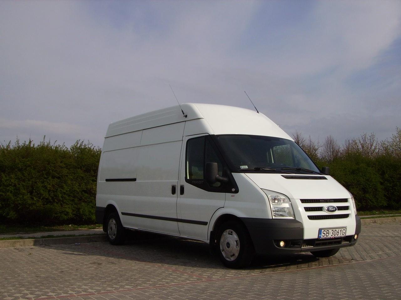 Biały, wysoki bus Ford Transit na kostce brukowej, z anteną na dachu i widoczną tablicą rejestracyjną, na tle zieleni i błękitnego nieba.