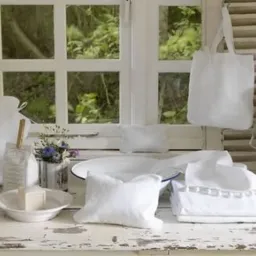Białe tekstylia domowe z lnu: poduszki, torba, termofor, ręczniki, mydło, prezentowane na rustykalnym, białym stole przed oknem z widokiem na zieleń.
