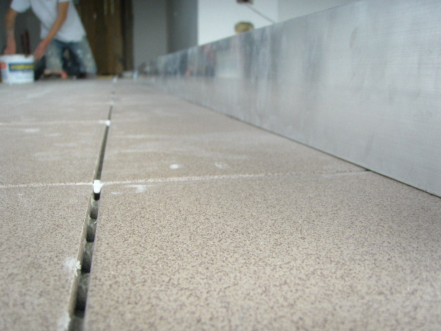 Układanie jasnych, kwadratowych płytek podłogowych z białymi krzyżykami dystansowymi, widoczna metalowa listwa przypodłogowa i osoba w tle z wiadrem kleju.