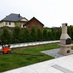 Pracownik w pomarańczowym kombinezonie używa maszyny do cięcia darni, przygotowując podłoże pod obrzeża trawnika, w tle widoczny murowany grill i rząd tuj.