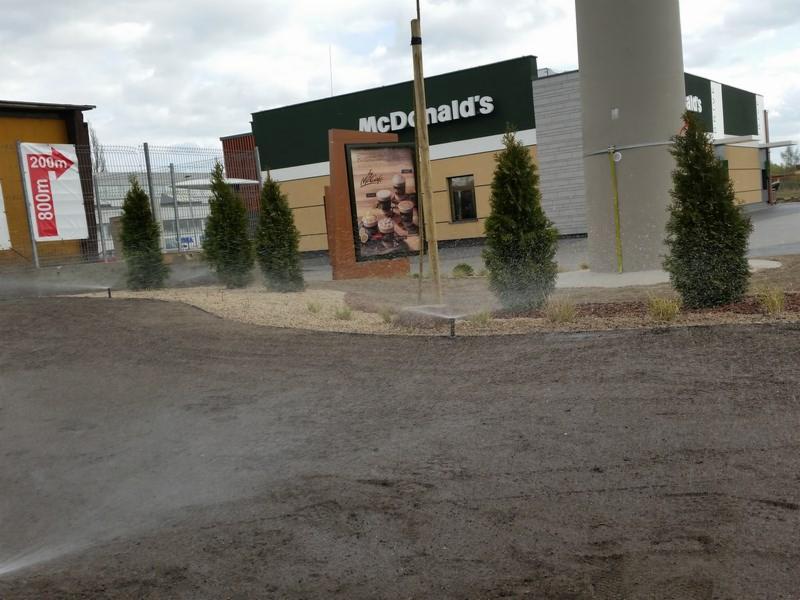 System nawadniania w Malborku w trakcie pracy, zraszający świeżo przygotowany teren pod trawnik, w tle rząd młodych drzewek iglastych i budynek restauracji.