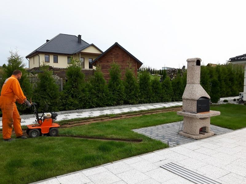 Pracownik w pomarańczowym kombinezonie używa maszyny do cięcia darni, przygotowując podłoże pod obrzeża trawnika, w tle widoczny murowany grill i rząd tuj.