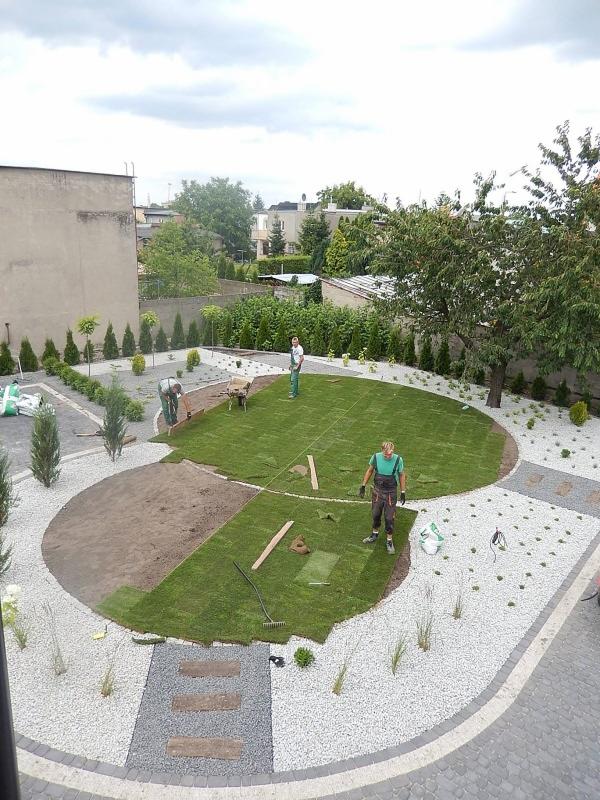 Układanie trawy z rolki w ogrodzie z wysypanym białym kamieniem, widoczne narzędzia i pracownicy w trakcie pracy.