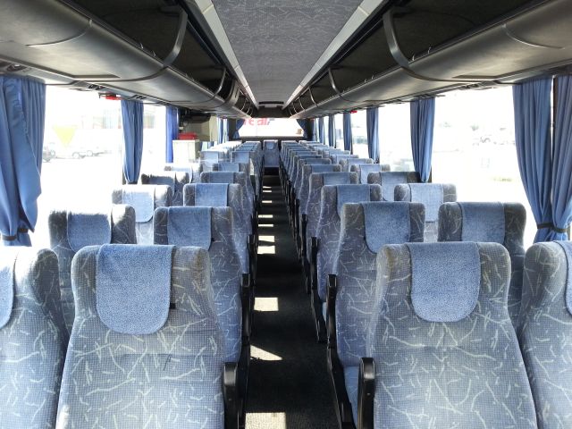 Wnętrze autobusu z rzędami niebieskich foteli z wzorem, zasłonięte okna po bokach, widok z przodu pojazdu.