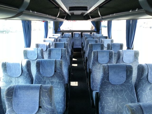 Wnętrze autokaru z rzędami niebieskich foteli z zagłówkami i zasłonami w oknach, widok z przodu pojazdu.