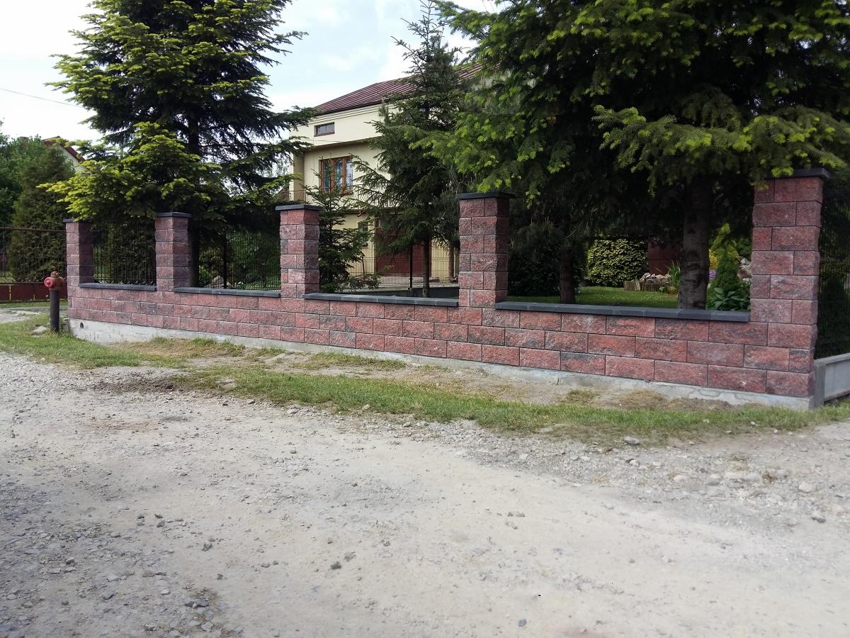 Nowoczesny, niski mur z czerwonej cegły klinkierowej z czarnymi elementami wykończeniowymi, tworzący ogrodzenie posesji z widocznym domem w tle, otoczony zielenią.