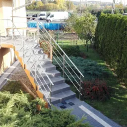 Zewnętrzne schody z szarymi płytkami i metalową balustradą z cienkich prętów, prowadzące do budynku. Widoczne krzewy i tuje w tle, oraz fragment elewacji budynku.