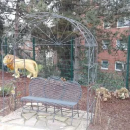 Metalowa konstrukcja łukowa z ławką i statuą lwa w ogrodzie. Zielone ogrodzenie siatkowe w tle. Elementy dekoracyjne i roślinność tworzą unikalny krajobraz.