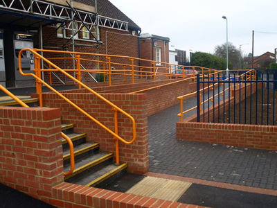 Zewnętrzne schody i rampa dla wózków inwalidzkich z pomarańczowymi poręczami Kee Klamp, wykonane z cegły, prowadzące do budynku z zadaszeniem.