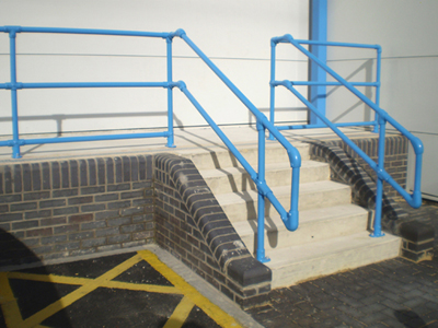 Zewnętrzne schody z betonowymi stopniami i niebieskimi, rurowymi poręczami Kee Klamp, otoczone ceglanym murkiem i chodnikiem z żółtymi oznaczeniami.