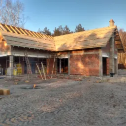 Surowa konstrukcja domu w trakcie budowy: murowane ściany z czerwonej cegły, drewniana konstrukcja dachu w trakcie montażu, rusztowania, teren budowy z piaskiem, ognisko i las w tle.