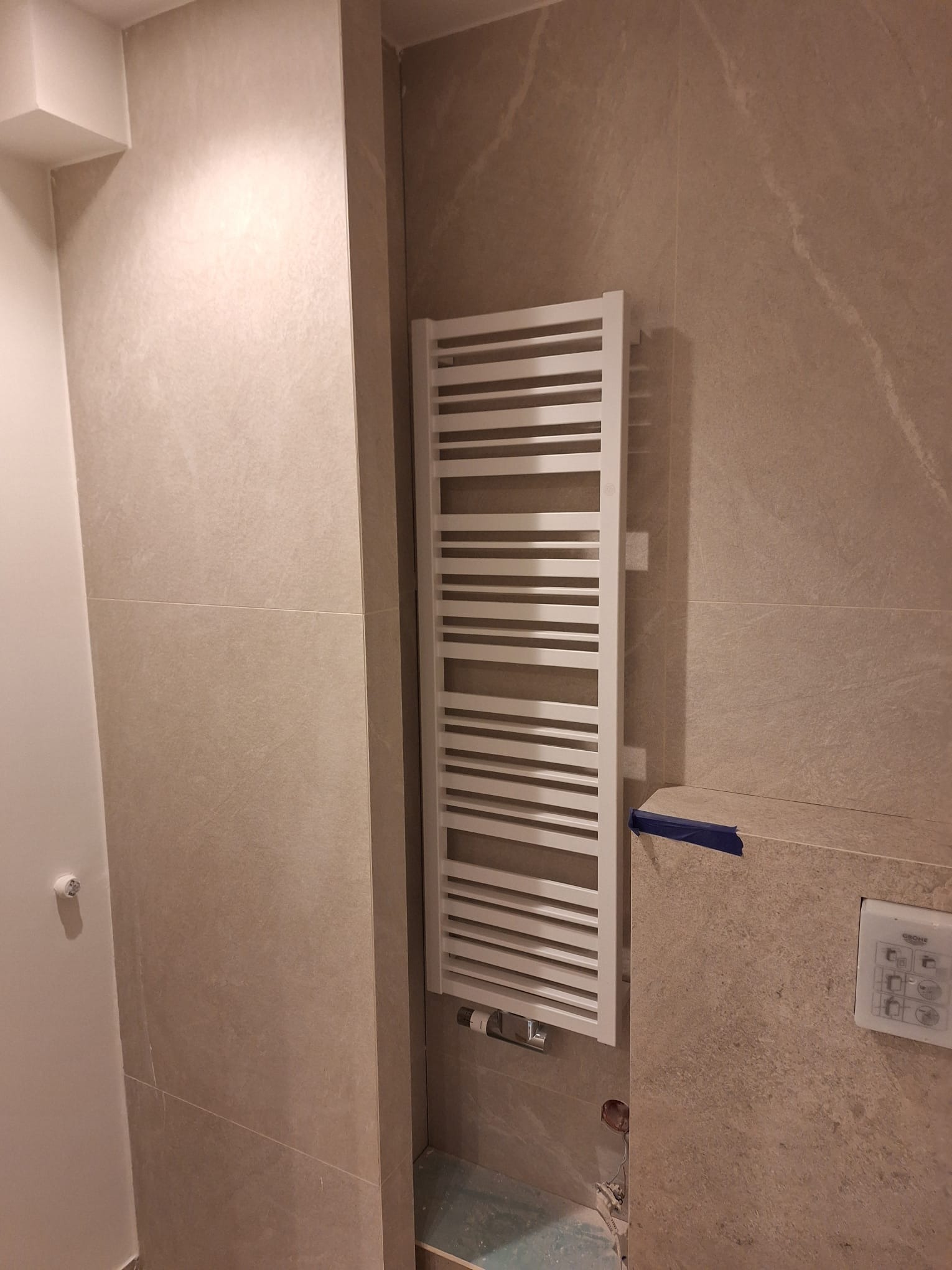 Montaż grzejnika drabinkowego w łazience wykończonej płytkami imitującymi kamień, widoczne niedokończone prace instalacyjne i zabezpieczenie taśmą malarską.