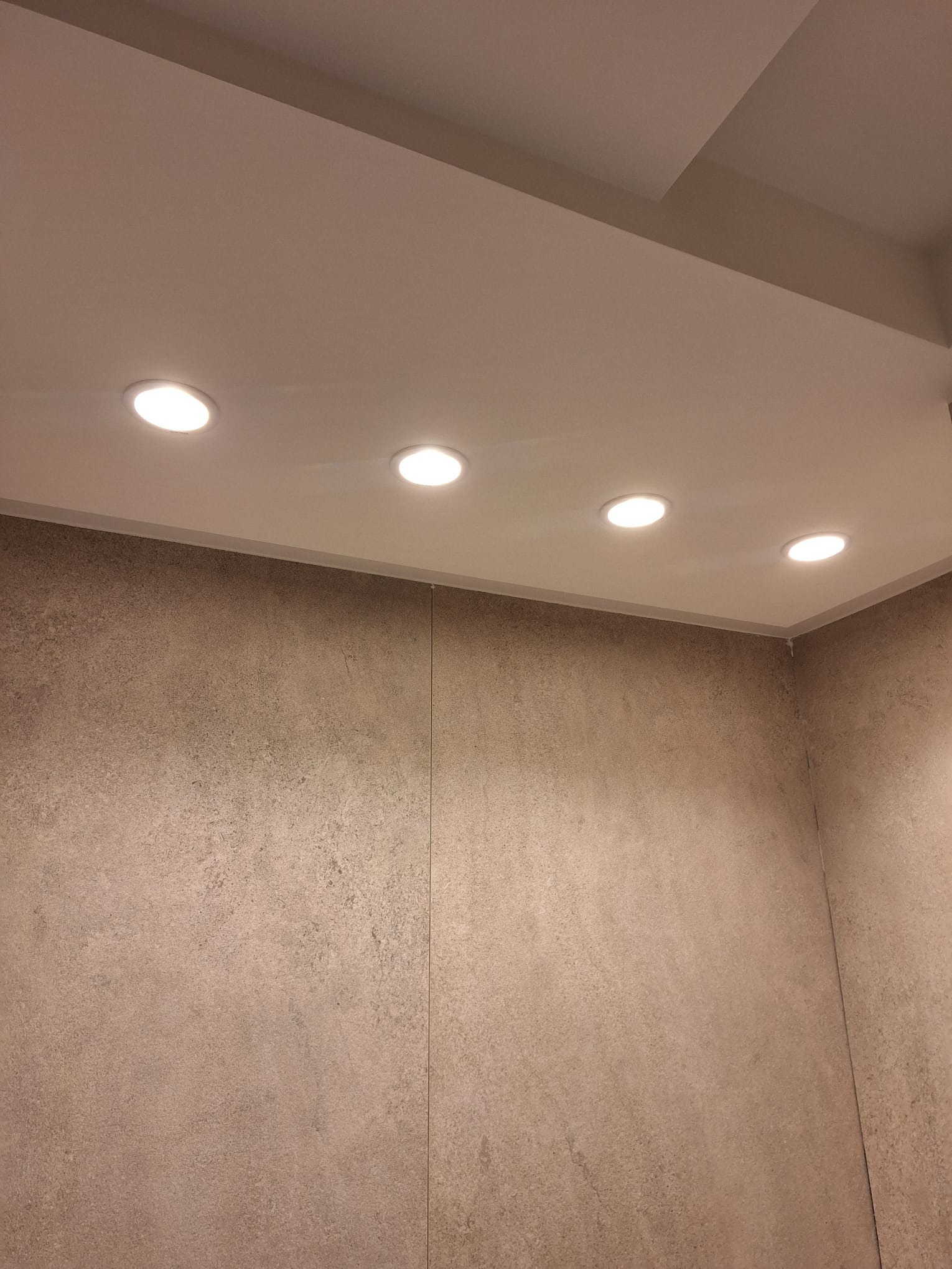 Wykończona łazienka z beżowymi płytkami imitującymi kamień na ścianach i sufitem podwieszanym z oświetleniem punktowym LED.