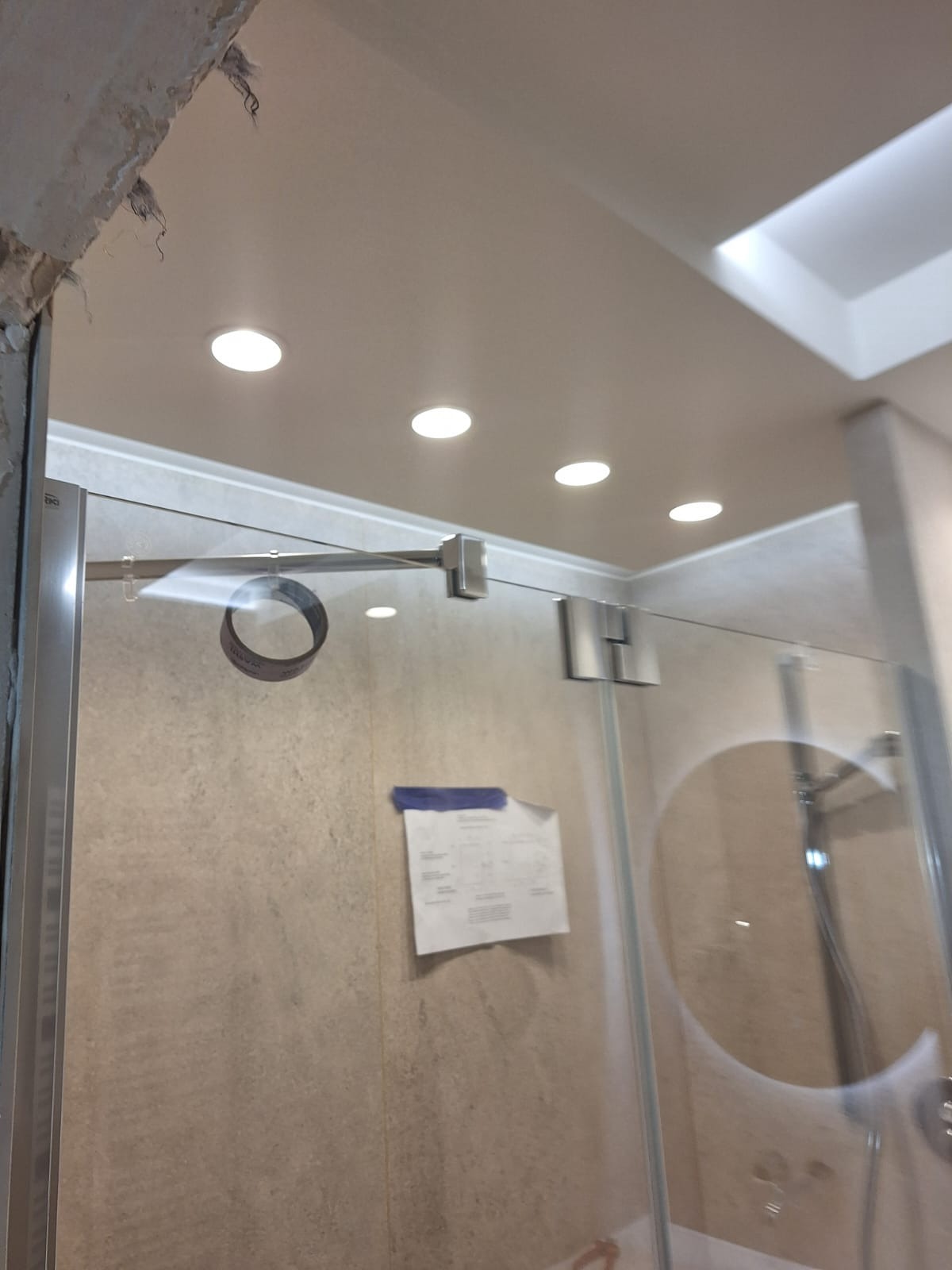 Szklana kabina prysznicowa z metalowymi okuciami, widoczne cztery okrągłe lampy w suficie, fragment ściany z niedokończonym wykończeniem i przyklejona kartka z rysunkiem technicznym.
