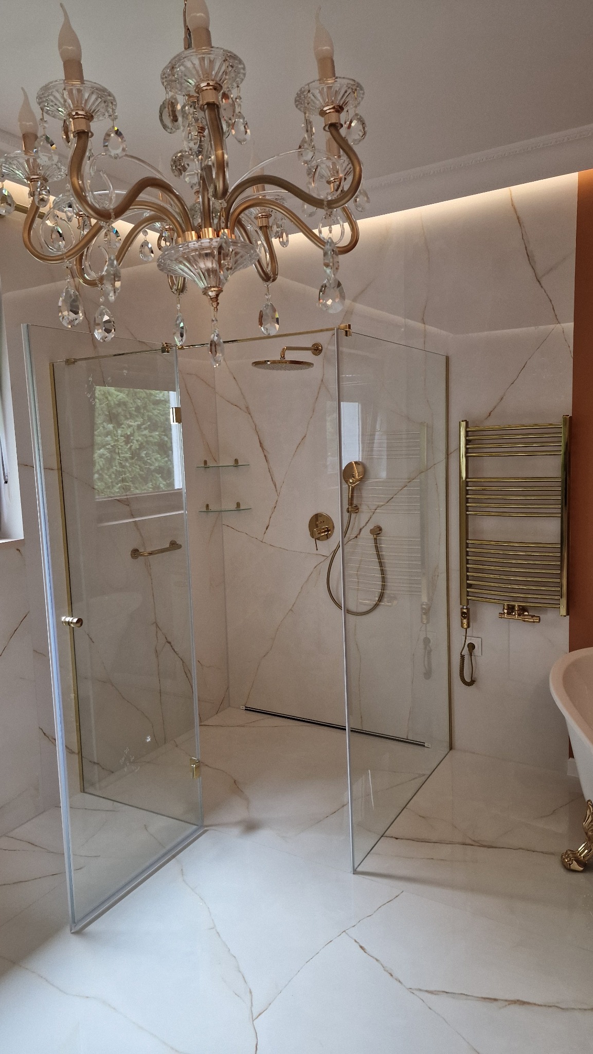 Luksusowa kabina prysznicowa z elementami złota, marmurowe płytki na podłodze i ścianach, ozdobny żyrandol kryształowy. Widok na elegancką łazienkę.