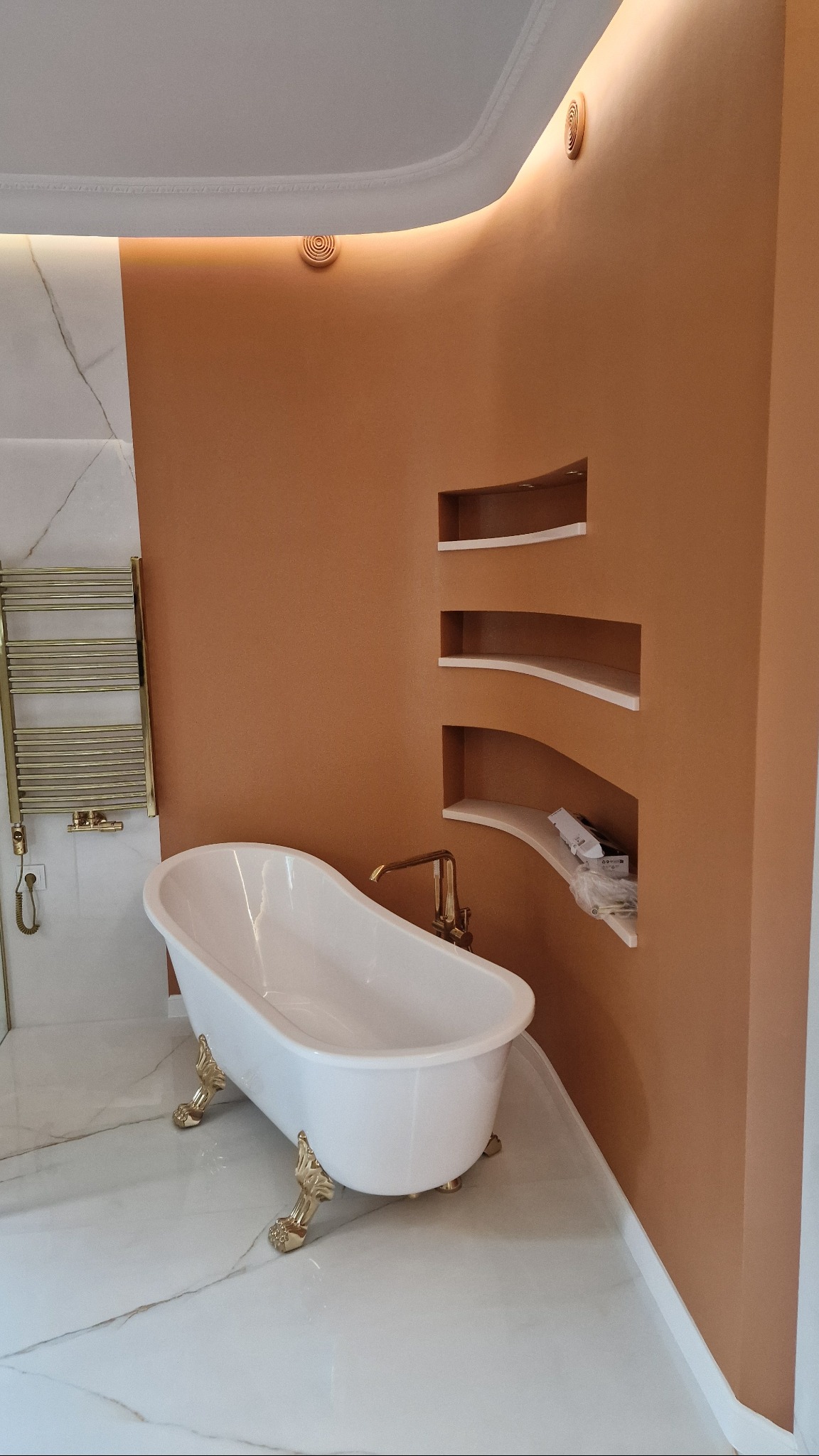 Elegancka łazienka z wolnostojącą wanną na złotych nóżkach, marmurową posadzką i ścianą w odcieniach brązu z wnękami. Widoczny złoty grzejnik i elementy wykończenia.