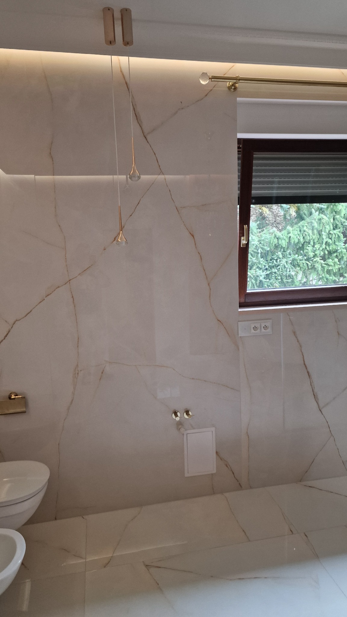 Elegancka łazienka wykończona płytkami imitującymi marmur z złotymi żyłami, wisząca lampa w kształcie kropli, okno z żaluzjami i armatura w złotym kolorze.