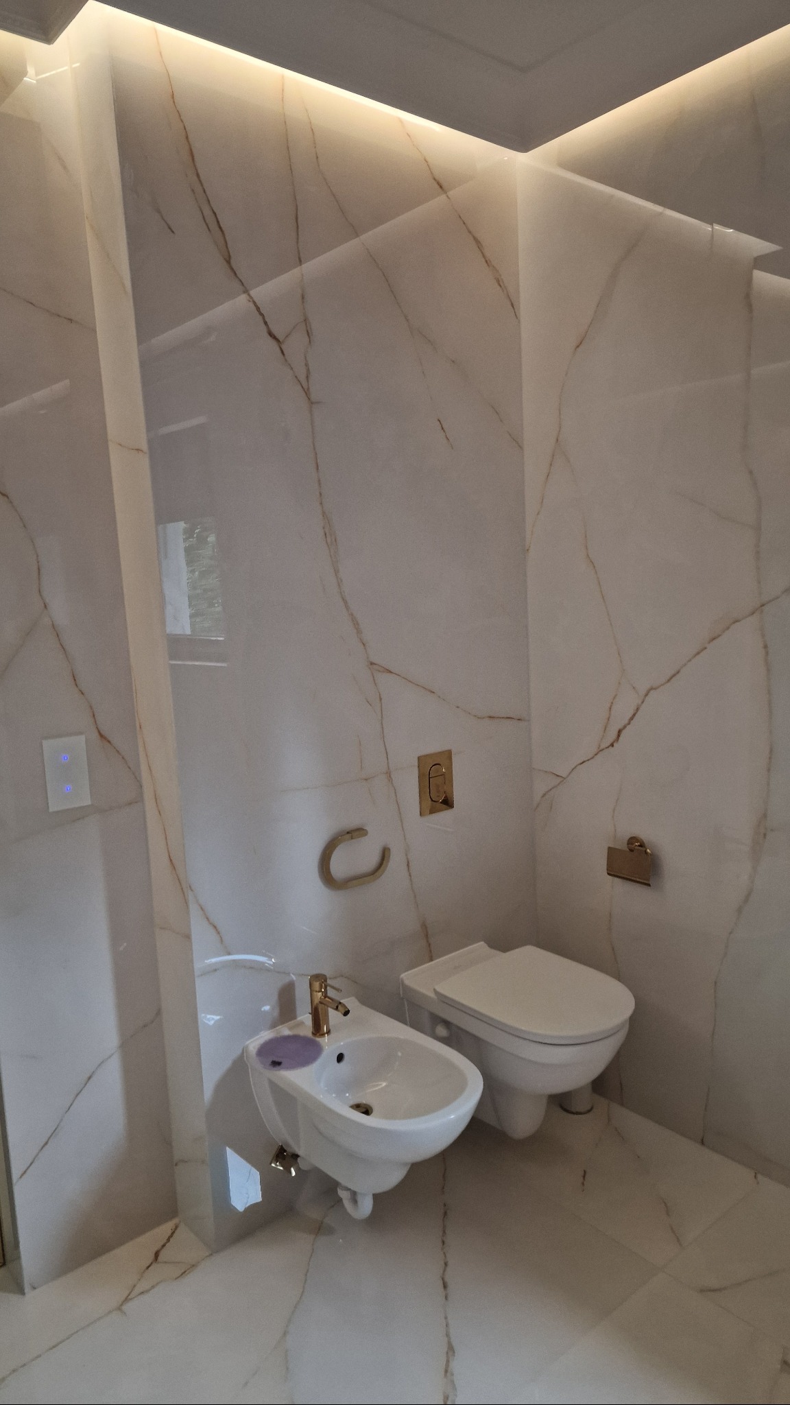 Elegancka łazienka wykończona płytami imitującymi marmur z oświetleniem LED, z wiszącą toaletą i bidetem z elementami w kolorze złota. Nowoczesny design.