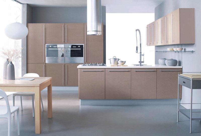 Nowoczesna kuchnia z drewnianymi frontami, wyposażona w szafki wiszące, piekarnik, płytę grzewczą i srebrny okap cylindryczny. Na blacie stoją garnki i miski, a obok znajduje się stół z krzesłami.